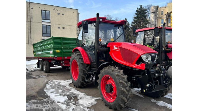 Na zdjęciu widać czerwony ciągnik rolniczy z podczepioną zieloną przyczepą, zaparkowany na placu z resztkami śniegu. Obok stoi drugi czerwony ciągnik. W tle są budynki i bezlistne drzewa, co wskazuje na zimę.